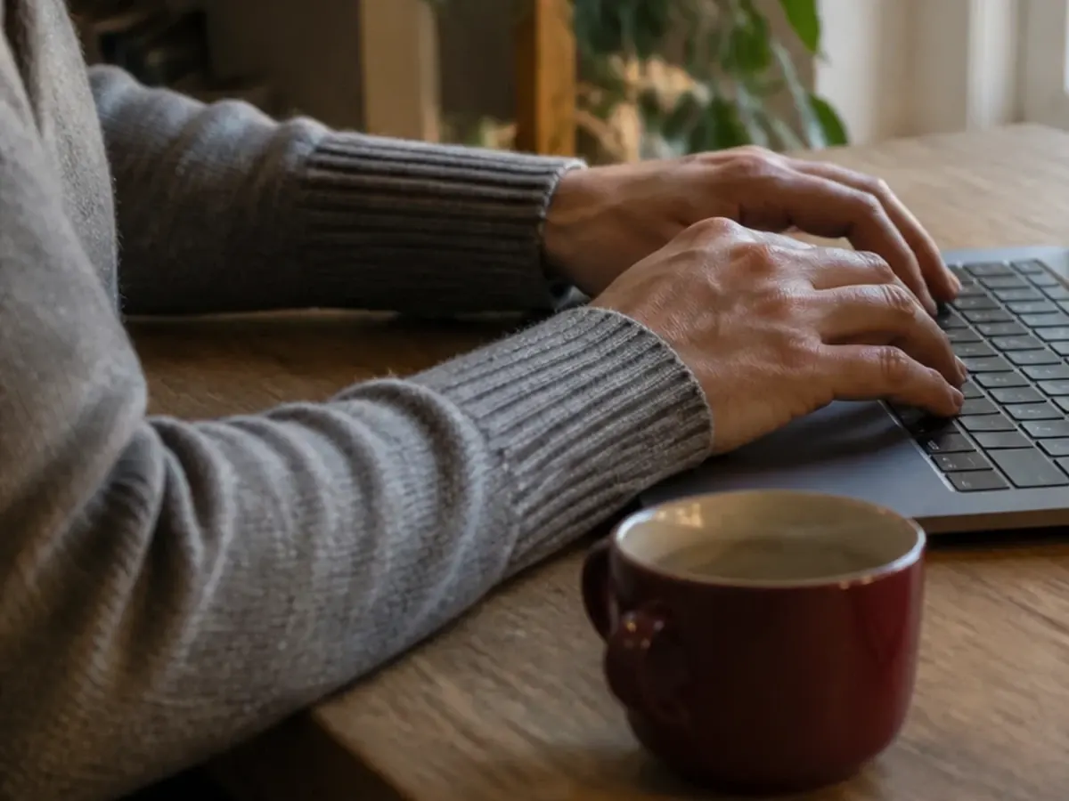 Manos con un jersey de punto escribiendo en el teclado de un portátil junto a una taza de café roja sobre una mesa de madera, con una planta detrás