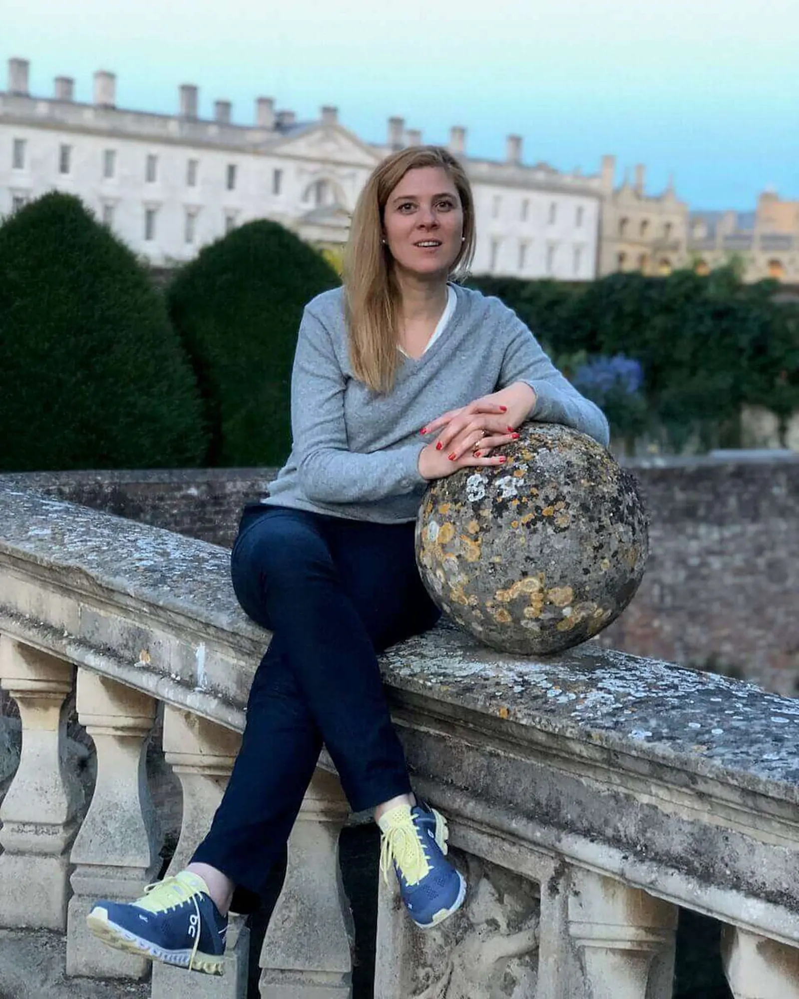Cecilia Altieri sitting on a stone balustrade at dusk, a country house and clipped hedges behind her