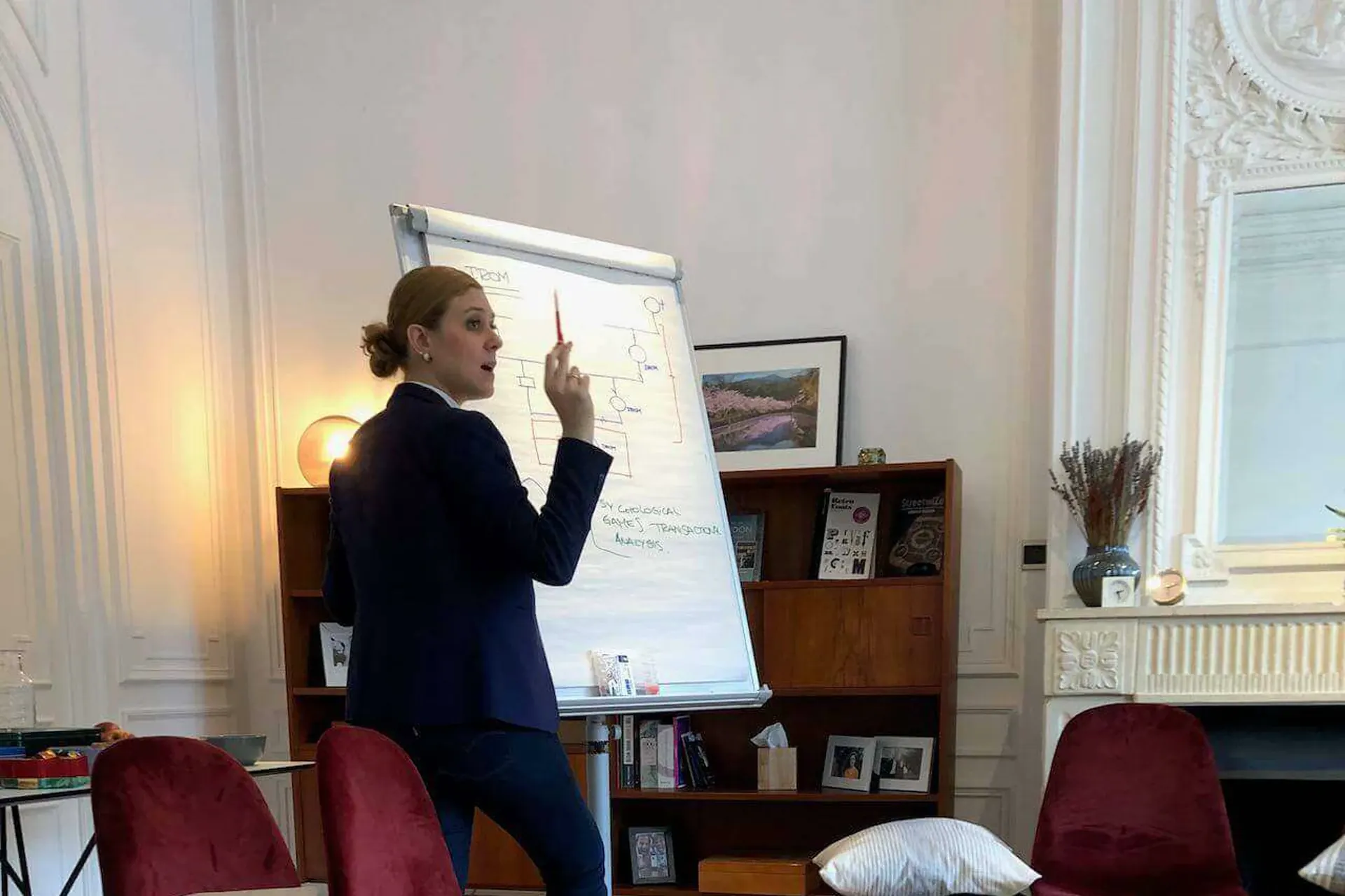 Cecilia junto a un rotafolio, con el rotulador rojo en alto, dirigiéndose a la sala a media frase junto a un genograma que ha dibujado